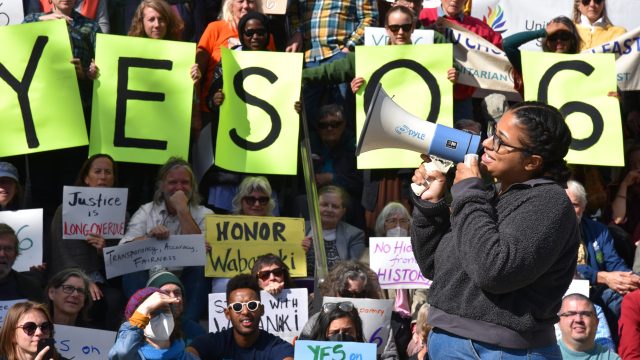 Reese Remington with Maine Votes/Maine Voices Network, a member of the Wabanaki Alliance Coalition, speaks to the crowd. Photo: Anne Henshaw Woman holding a megaphone speaking to crowd with people holding yellow signs with letters Y E S Q 6