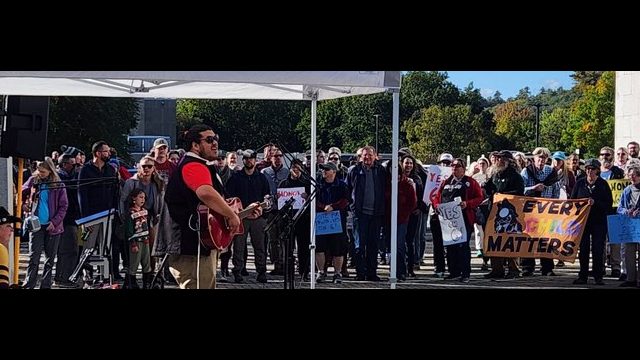 Just Honey of Honey Buzz Studios in Penobscot County performed for a crowd of more than 200. Photo: Rob Laraway Person playing guitar and singing in front of large crowd for Indigenous Peoples' Day Rally held by Wabanaki Alliance
