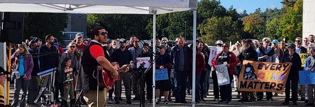 Just Honey of Honey Buzz Studios in Penobscot County performed for a crowd of more than 200. Photo: Rob Laraway Person playing guitar and singing in front of large crowd for Indigenous Peoples' Day Rally held by Wabanaki Alliance