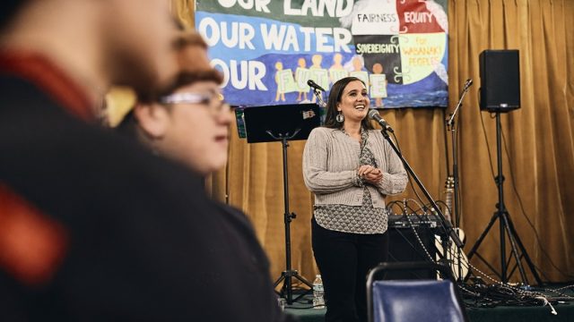 Maulian Bryant, Penobscot Nation Ambassador and President of the Wabanaki Alliance, welcomed guests to Rising Voices. Maulian Bryant, Penobscot Nation Ambassador and President of the Wabanaki Alliance, speaks at a microphone, smiling, hands clasped, with colorful banner in background with Our Land, Our Water, Our Future