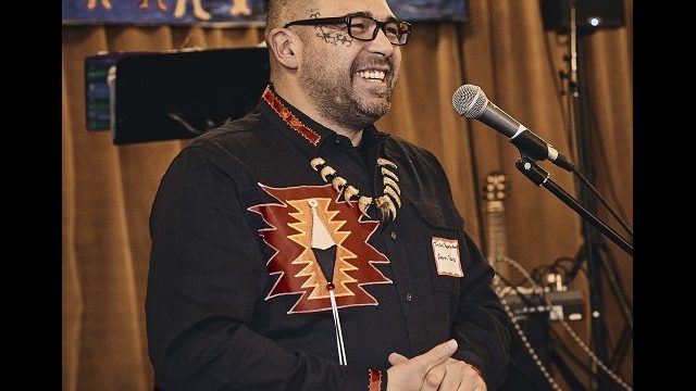 Aaron Dana, Passamaquoddy Tribal Representative in the Maine House of Representatives, speaks at the event. Aaron Dana, Passamaquoddy Tribal Representative in the Maine House of Representatives stands at a microphone smiling, hands clasped